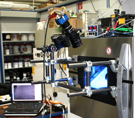 Camera setup in front of LBM machine (EOSINT M 270, EOS GmbH, Germany). The modular tube construction enables flexible positioning with adjustable height, position, and distance from door. A geared head allows three-axis rotation of camera and camera lens to align it with the build platform.